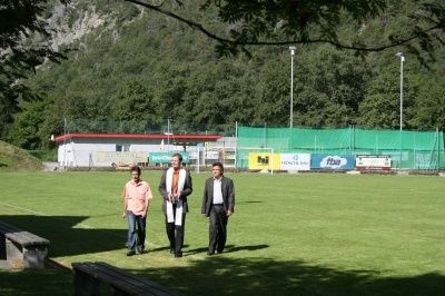 Drei Männer gehen über ein Grasfeld, mit einem Gebäude und Bäumen im Hintergrund. Ein Mann hält ein weißes Tuch.