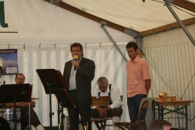 Ein Mann steht vor einem Mikrofon, möglicherweise singend, mit anderen sitzend und stehend um ihn herum in einem Zelt.