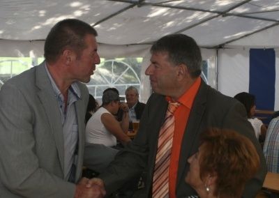 Zwei Männer in Anzügen schütteln sich die Hände in einem Zelt, mit einer Frau und anderen Personen im Hintergrund.