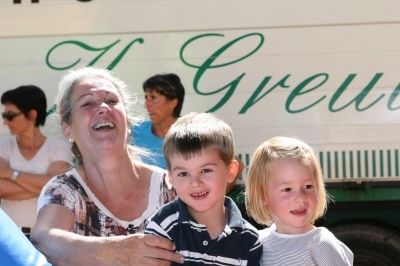 Eine Frau mit grauen Haaren hält zwei Kinder, einen Jungen und ein Mädchen, vor einem weißen Bus mit grüner Schrift. Die Frau lächelt und scheint zu lachen. Die Kinder schauen in die Kamera. Dahinter stehen zwei Frauen, von denen eine eine Sonnenbrille trägt.