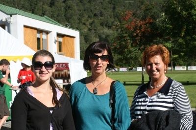 Drei Frauen stehen vor einem Gebäude, eine mit Sonnenbrille und eine mit einer Halskette. Sie lächeln und posieren für ein Foto. Dahinter ist ein Rasenfeld und Bäume zu sehen.