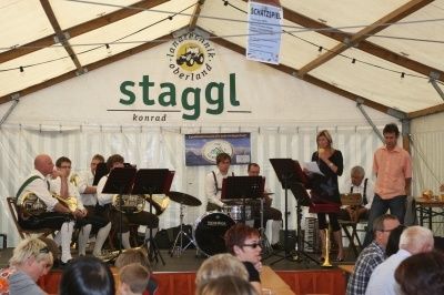 Eine Band spielt auf der Bühne in einem Zelt mit dem Schriftzug 'Stagggl Konrad'. Das Publikum sitzt davor, einige tragen Hüte und Brillen. Musikinstrumente stehen auf der Bühne, darunter Trommeln, Trompeten und ein Posaune.