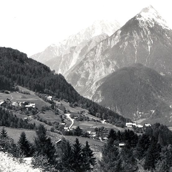 Ein Schwarz-Weiß-Foto einer Berglandschaft mit einem kleinen Dorf im Tal darunter.