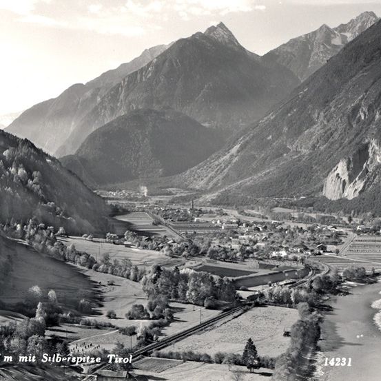 Eine Schwarz-Weiß-Luftaufnahme eines Tals mit Bergen, einem Fluss und einer Stadt. Das Tal ist von Bäumen und Bergen umgeben.