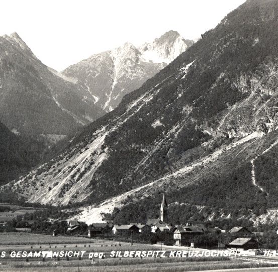 Ein Schwarz-Weiß-Foto einer Berglandschaft mit einem Dorf darunter, mit einem Kirchturm und einer Eisenbahnstrecke.
