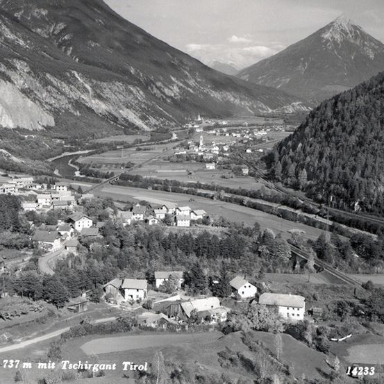 Eine schwarz-weiße Luftaufnahme eines Dorfes, das zwischen Bergen und einem Fluss eingebettet ist. Das Dorf ist von üppigem Grün umgeben und hat eine Mischung aus traditionellen und modernen Gebäuden.