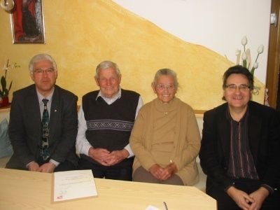 Vier Personen sitzen an einem Tisch und lächeln für ein Foto. Eine Frau in der Mitte hält einen Stift. Ein gerahmtes Bild und Blumen schmücken die Wand hinter ihnen.