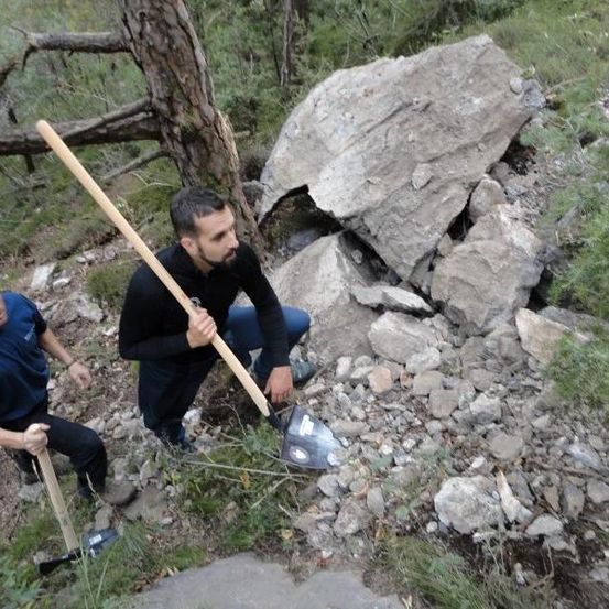 Zwei Männer graben mit Schaufeln in einem felsigen Bereich, einer in einem blauen Shirt und der andere in Schwarz.