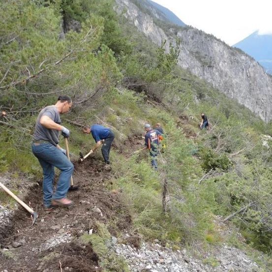 Eine Gruppe von Menschen arbeitet auf einem steilen Hang und benutzt Schaufeln und Rechen. Sie sind in Freizeitkleidung gekleidet und scheinen an einer Art Instandhaltungs- oder Bauarbeit beteiligt zu sein.