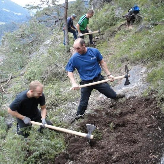 Vier Männer arbeiten auf einem Hang und benutzen Äxte, um den Boden zu graben und zu räumen. Der Mann im blauen Shirt hält eine Axt und scheint die Gruppe zu führen.