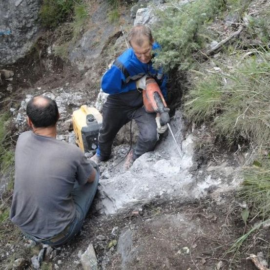 Zwei Männer arbeiten auf einem felsigen Hang, einer bohrt mit einem Bohrer in den Felsen, während der andere zusieht.