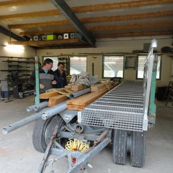 Zwei Männer stehen neben einem Anhänger, der mit Holzbrettern und Metallrohren beladen ist, in einer Werkstatt.