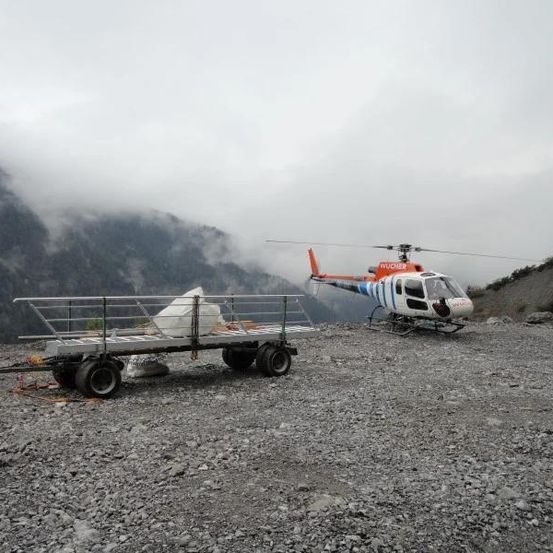 Ein Hubschrauber ist auf einer felsigen Oberfläche geparkt, in der Nähe ein Frachtanhänger, Berge im Hintergrund.