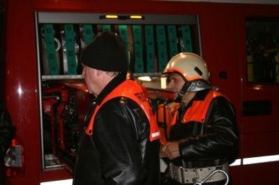 Zwei Feuerwehrleute in voller Ausrüstung stehen vor einem Feuerwehrwagen, einer scheint Anweisungen zu geben.