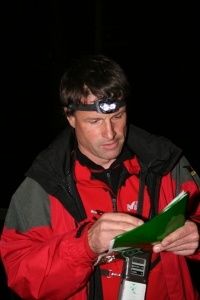 Ein Mann in einem roten Jackett mit Stirnlampe schaut in einem dunklen Bereich in ein grünes Notizbuch.