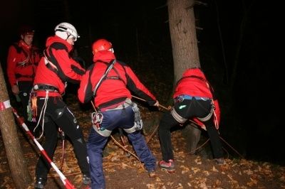 Vier Personen in roten Jacken und Helmen führen nachts in einem Wald eine Rettungsaktion durch und helfen jemandem mit Seilen.