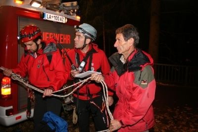 Drei Männer in roten Jacken und Helmen stehen neben einem Feuerwehrauto und halten ein Seil. Sie scheinen etwas zu besprechen.