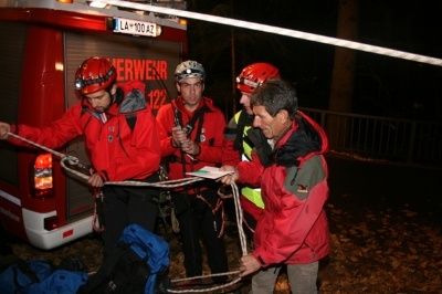 Fünf Personen in roten und gelben Jacken stehen um einen Feuerwehrwagen und halten Seile, während sie über eine Rettungsaktion sprechen.