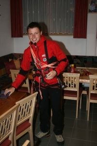 Ein Mann in einem roten Jackett und schwarzen Hosen steht in einem Esszimmer und hält Kletterausrüstung. Hinter ihm ist ein Tisch mit Stühlen und ein roter Vorhang zu sehen.
