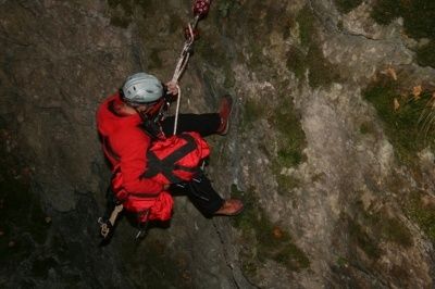 Eine Person in einem roten Jackett und Helm klettert mit einem Seil an einer steilen Felswand hinunter.