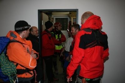 Eine Gruppe von Menschen in orangen und roten Jacken versammelt sich in einem Raum, möglicherweise zur Vorbereitung einer Outdoor-Aktivität. Einige tragen Helme und Rucksäcke.