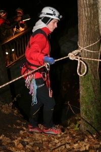 Eine Person in einem roten Jackett und Helm klettert mit Seilen und Handschuhen an einem Baum. Die Szene ist dunkel, und es gibt Blätter auf dem Boden.