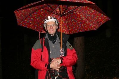 Ein Mann in einem roten Mantel und Helm steht in einem dunklen Wald und hält einen rot gepunkteten Regenschirm. Er scheint auf etwas oder jemanden zu warten.