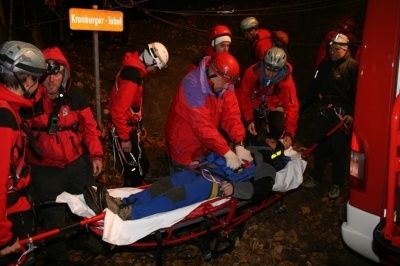 Eine Gruppe von Menschen in roten Jacken und Helmen trägt eine Trage mit einer Person darauf. Sie befinden sich nachts in einem Wald.