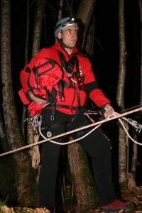 Ein Mann in einem roten Jackett und Helm steht nachts in einem Wald und hält ein Seil. Er ist mit Kletterausrüstung ausgestattet und scheint sich auf eine Aktivität vorzubereiten.