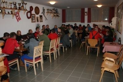 Eine Gruppe von Menschen sitzt an Tischen in einem Restaurant, einige tragen rote Jacken, und es gibt Trophäen an der Wand.