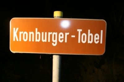 Ein orangefarbenes Straßenschild mit weißem Text 'Kronburger-Tobel' ist nachts an einem Pfosten beleuchtet.