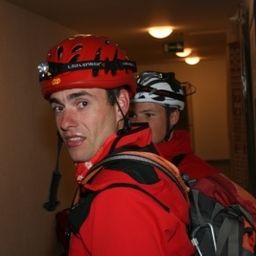 Zwei Personen in roten Jacken und Helmen stehen in einem Flur, einer trägt einen Rucksack.