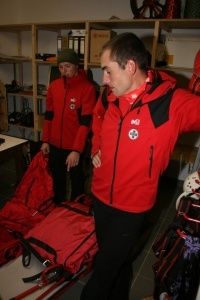 Zwei Männer in roten Jacken mit roten Taschen und Skiausrüstung in einem Raum mit Regalen und einem weißen Tisch.
