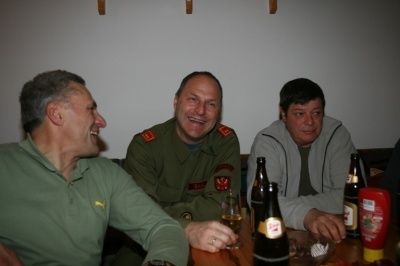 Drei Männer sitzen an einem Tisch, lächeln und trinken. Einer trägt eine Militäruniform, ein anderer ein grünes Hemd und der dritte eine graue Kapuzenjacke. Flaschen und Gläser stehen auf dem Tisch.