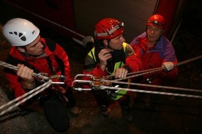 Drei Personen in roten Jacken und Helmen sind in eine Seilaktivität verwickelt, möglicherweise eine Rettungsaktion. Einer hält ein Seil, ein anderer spricht in ein Funkgerät und der dritte hält ein Seil mit einem Metallgerät.