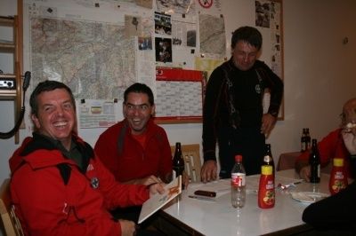 Vier Männer in roten Jacken sitzen an einem Tisch mit verschiedenen Flaschen und Gegenständen. Ein Mann hält ein Stück Papier und lächelt. Hinter ihnen ist eine Wand mit einer Karte und anderen Papieren.