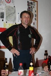 Ein Mann in einem schwarzen Langarmshirt mit rotem Rand und Hosenträger steht an einem Tisch mit mehreren Flaschen und einer Plastikflasche. Er hat die Hände auf den Hüften und lächelt. Hinter ihm hängen Poster an der Wand.