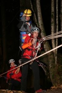 Drei Personen in roten Jacken und Helmen führen nachts eine Rettungsaktion in einem Wald durch, wobei eine Person eine Seilleiter erklimmt, während die anderen helfen.