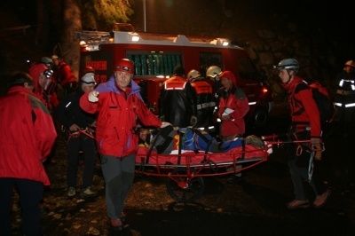 Eine Gruppe von Rettungskräften in roten Jacken und Helmen trägt eine Trage mit einer Person bei Nacht. Ein Feuerwehrauto steht hinter ihnen.