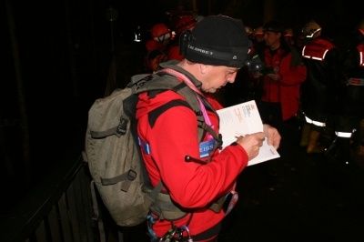 Ein Mann in einem roten Jacke und schwarze Mütze schreibt auf ein Stück Papier, während er einen Rucksack trägt und in einem schwach beleuchteten Bereich steht, in dem sich andere Personen in roten Jacken befinden.