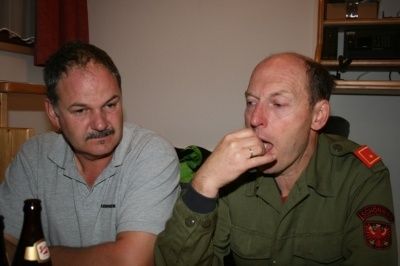 Zwei Männer sitzen in einem Raum, einer in einem grauen Hemd und der andere in einer grünen Militäruniform, letzterer pustet sich die Nase.