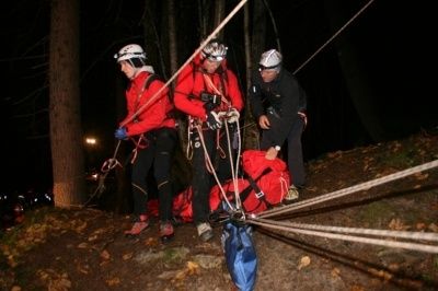 Drei Personen in roten Jacken und Helmen sichern Seile an einem steilen, bewaldeten Hang bei Nacht. Eine Person hält eine rote Tasche, während eine andere eine blaue Tasche hält.