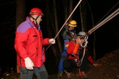 Drei Personen in Helmen und Sicherheitsausrüstung sind in einem Waldgebiet an einer nächtlichen Rettungsaktion beteiligt. Eine Person hält ein Seil, eine andere kniet, und die dritte steht.