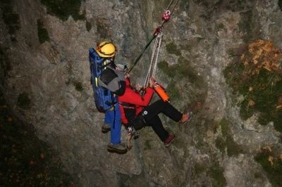 Zwei Personen werden nachts von einem steilen Fels gerettet. Eine Person trägt eine rote Jacke und wird von einem Seil hochgezogen. Die andere Person trägt einen gelben Helm und einen blauen Rucksack.