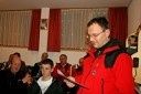 Ein Mann in einem roten Mantel steht vor einer Gruppe von Menschen und liest laut aus einem Stück Papier.