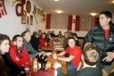 Eine Gruppe von Menschen in roten Shirts sitzt um einen Tisch in einem Restaurant, mit Flaschen und Gläsern auf dem Tisch. Ein Mann in einer schwarzen Jacke steht und spricht mit ihnen.