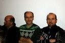 Vier Männer stehen zusammen und lächeln für ein Foto. Der Mann in der Mitte trägt einen grün-weiß gemusterten Pullover, während die anderen schwarze Jacken tragen. Sie befinden sich in einem Raum mit einer weißen Wand.