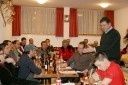 Eine Gruppe von Menschen sitzt um einen Tisch in einem Esszimmer, mit einem Mann, der vorne steht und möglicherweise eine Rede oder Präsentation hält. Der Raum hat rote Vorhänge und eine warme Beleuchtung.