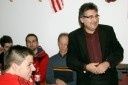 Ein Mann in einem schwarzen Anzug steht vor einer Gruppe von Menschen, lächelt und gestikuliert mit den Händen. Hinter ihm sitzen drei Männer in Stühlen, einer hält eine Mappe. Eine Flagge hängt an der Wand.