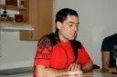 Ein Mann mit langen Haaren, der ein rotes Shirt trägt, sitzt an einem Tisch und schaut nach unten. Hinter ihm befindet sich ein Holzschrank mit Regalen und einer weißen Wand. Rechts neben ihm sitzt eine weitere Person.